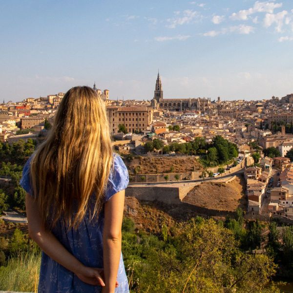 A woman with long blonde hair stands overlooking a historic cityscape under a clear blue sky.