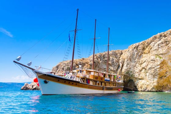A large wooden sailboat with multiple masts is anchored near a rocky cliff under a clear blue sky.
