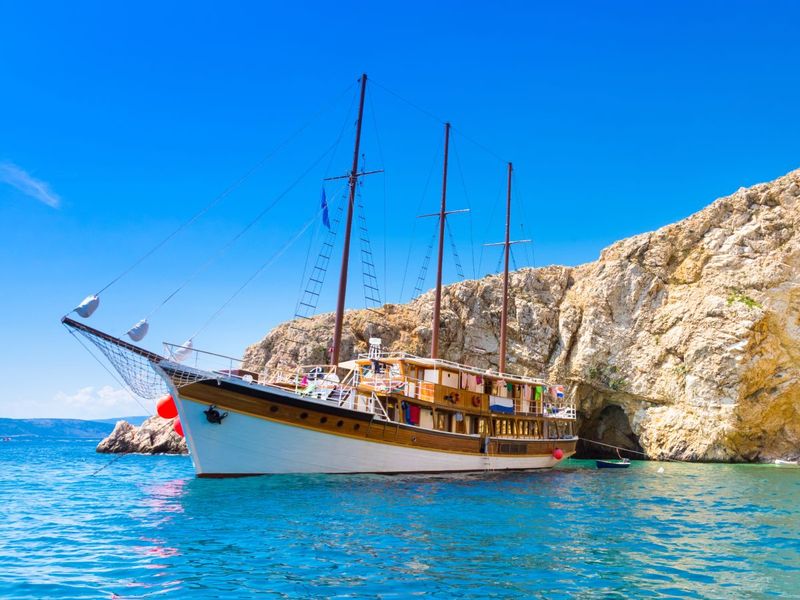 A large wooden sailboat with multiple masts is anchored near a rocky cliff under a clear blue sky.