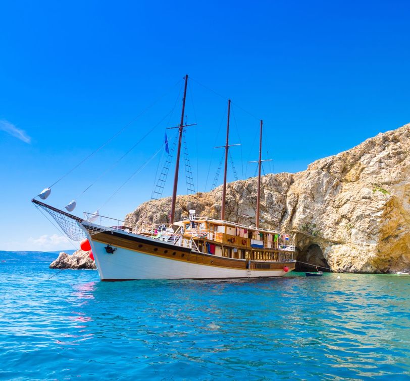 A large wooden sailboat with multiple masts is anchored near a rocky cliff under a clear blue sky.