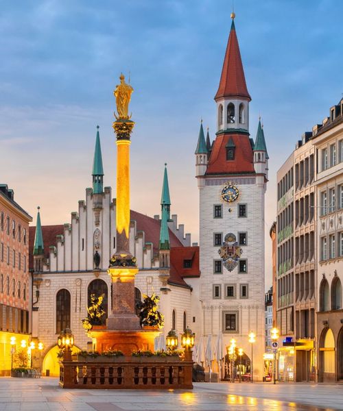 A historic square with ornate buildings and a tall column topped with a golden statue, illuminated at dusk in a European city.