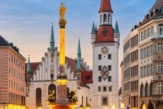 A historic square with ornate buildings and a tall column topped with a golden statue, illuminated at dusk in a European city.