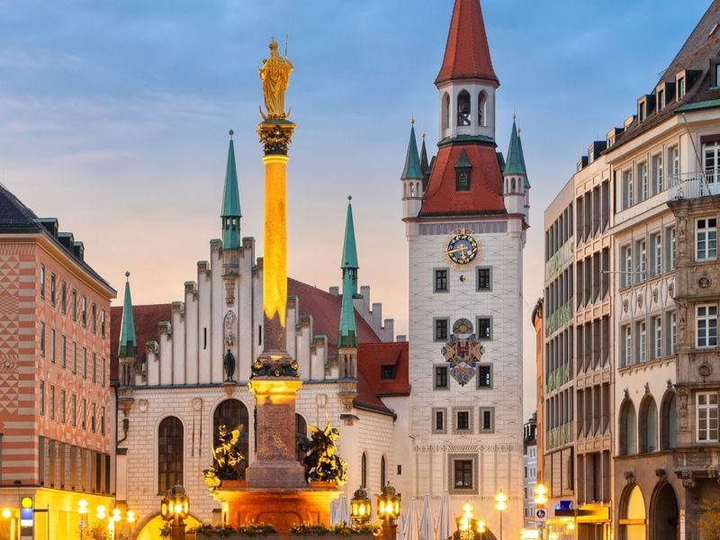 A historic square with ornate buildings and a tall column topped with a golden statue, illuminated at dusk in a European city.