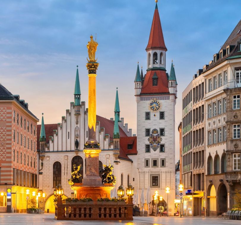 A historic square with ornate buildings and a tall column topped with a golden statue, illuminated at dusk in a European city.