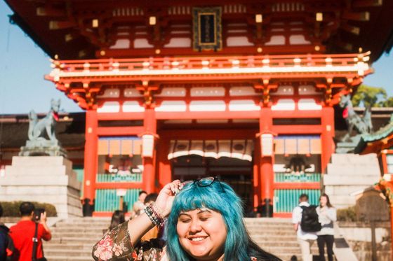 A smiling person with blue hair stands in front of a vibrant red temple, wearing a floral jacket and holding sunglasses.