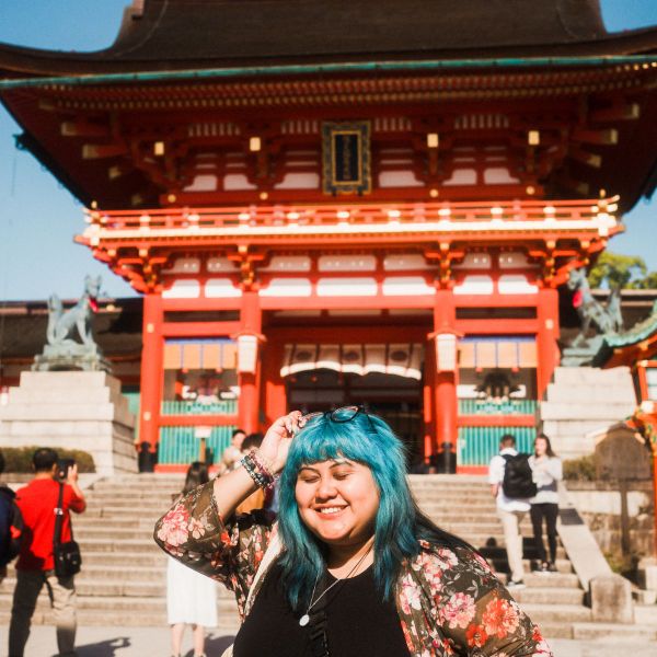 A smiling person with blue hair stands in front of a vibrant red temple, wearing a floral jacket and holding sunglasses.