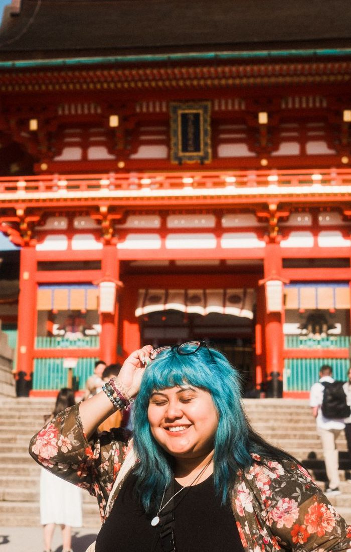 A smiling person with blue hair stands in front of a vibrant red temple, wearing a floral jacket and holding sunglasses.