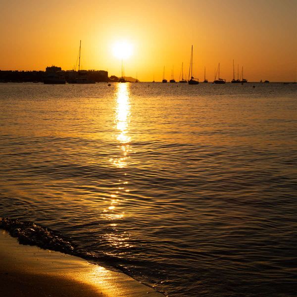 Sunset over a calm sea with silhouetted sailboats and a golden reflection on the water, near a sandy shore.