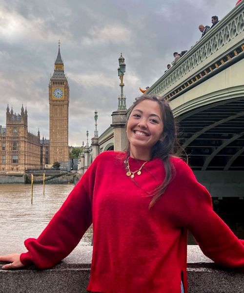 Smiling person in a red sweater, standing by a river with Big Ben and a bridge in the background.