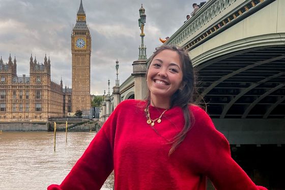 Smiling person in a red sweater, standing by a river with Big Ben and a bridge in the background.