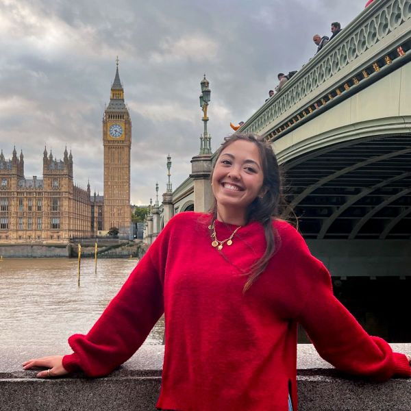 Smiling person in a red sweater, standing by a river with Big Ben and a bridge in the background.