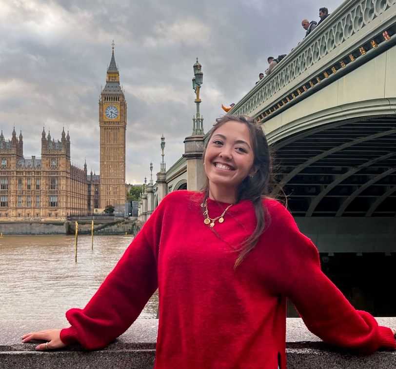 Smiling person in a red sweater, standing by a river with Big Ben and a bridge in the background.