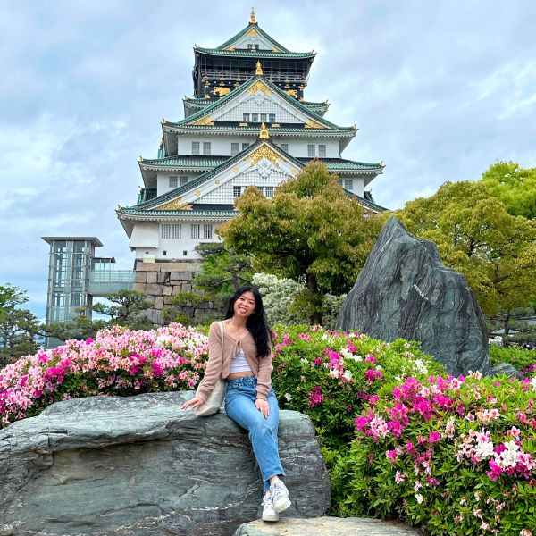 A woman sits on a rock surrounded by pink flowers in front of Osaka Castle, with cloudy skies above.