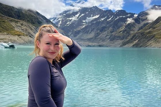 Woman smiling by a turquoise lake with mountains in the background, holding a camera and shielding her eyes from the sun.