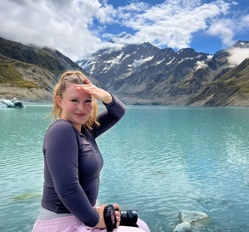 Woman smiling by a turquoise lake with mountains in the background, holding a camera and shielding her eyes from the sun.