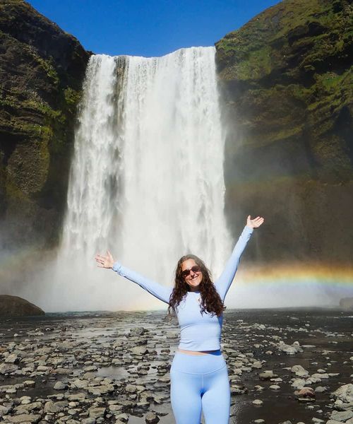 A person in light blue attire stands with arms raised in front of a tall waterfall, with a rainbow visible in the mist.