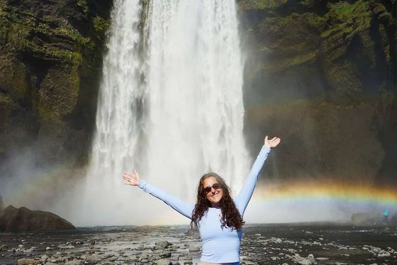 A person in light blue attire stands with arms raised in front of a tall waterfall, with a rainbow visible in the mist.