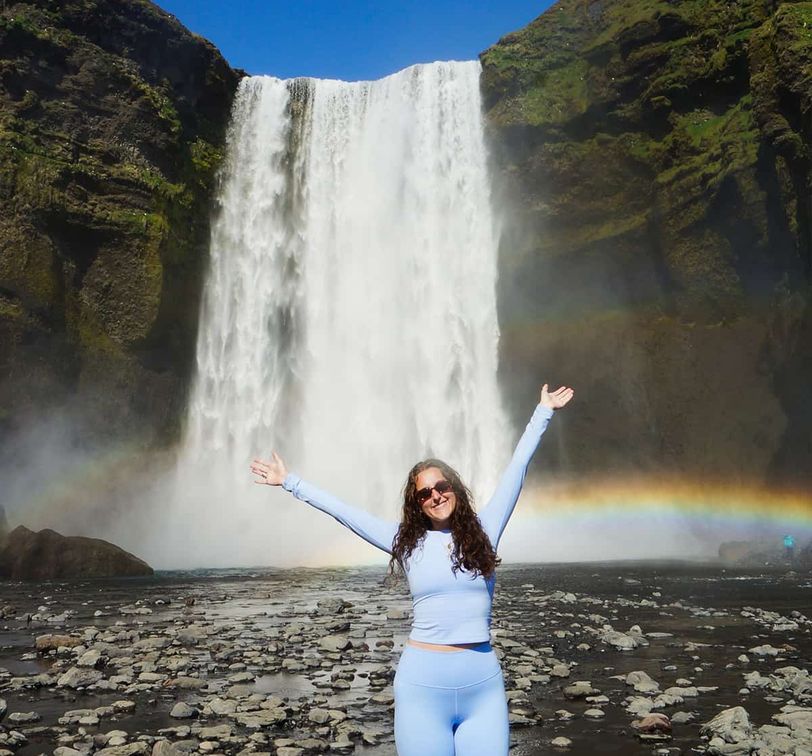 A woman in light blue clothing stands with arms raised in front of a large waterfall with a rainbow, surrounded by rocky terrain.