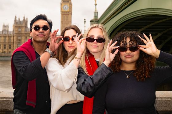 Four people wearing sunglasses and posing playfully near Big Ben and Westminster Bridge on a cloudy day.