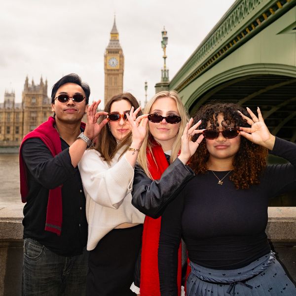 Four people wearing sunglasses and posing playfully near Big Ben and Westminster Bridge on a cloudy day.