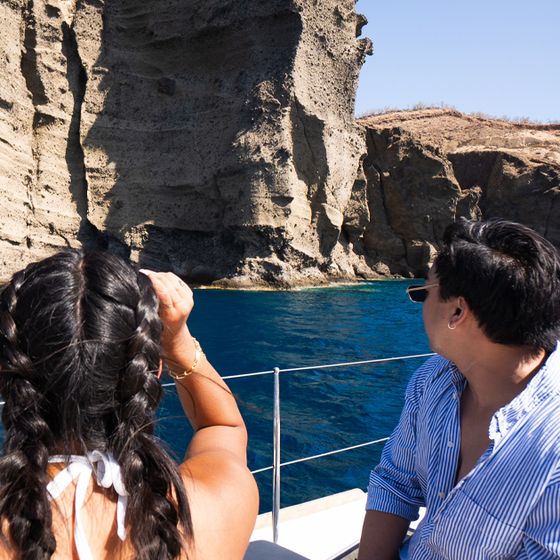 A couple on a boat looks at a rocky cliff over clear blue water under a clear sky. The woman has braided hair; the man wears sunglasses and a striped shirt.