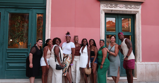 A group of twelve people smiling and posing in front of a pink building with green doors and windows.