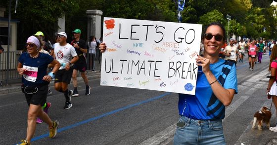 A smiling woman holds a handmade sign supporting runners in a race. Runners pass by on the street, with trees and spectators in the background.