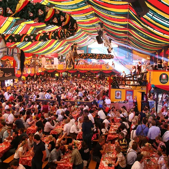 Crowded festival tent with colorful drapes, chandeliers, and people wearing traditional attire, enjoying food and drinks at long tables.