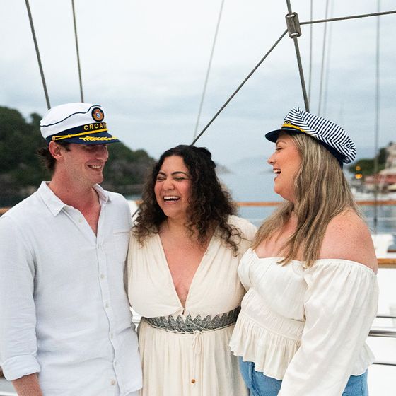 Three people wearing sailor hats laugh together on a boat, with trees and a building visible in the background.