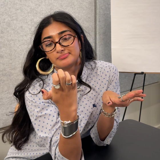 A woman with glasses and long hair examines her nails while sitting at a table, wearing large hoop earrings and multiple silver rings.