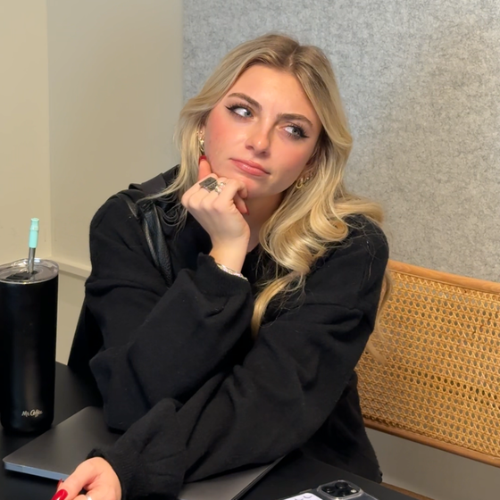 A woman with long blonde hair sits at a desk, resting her chin on her hand and looking thoughtfully to the side.