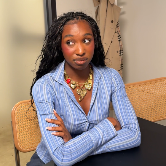 Woman with long braided hair and striped blue shirt sits at a table with arms crossed, wearing a statement necklace, looking up thoughtfully.
