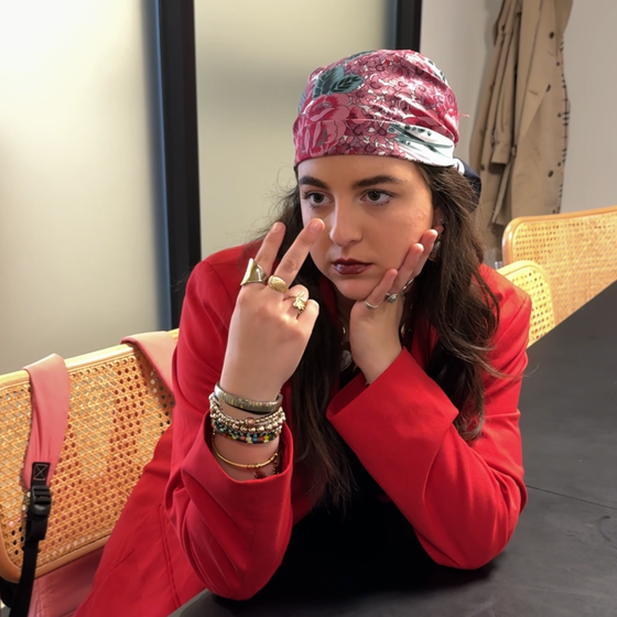 A woman in a red outfit and patterned headscarf rests her head on her hand while gesturing with two fingers, seated at a table.
