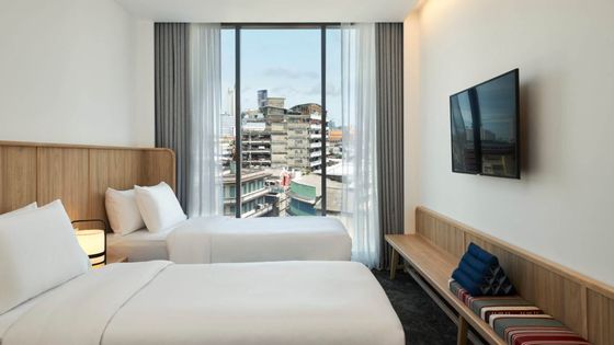 Modern hotel room with two beds, large window overlooking city buildings, wall-mounted TV, and a bench with cushions beneath.