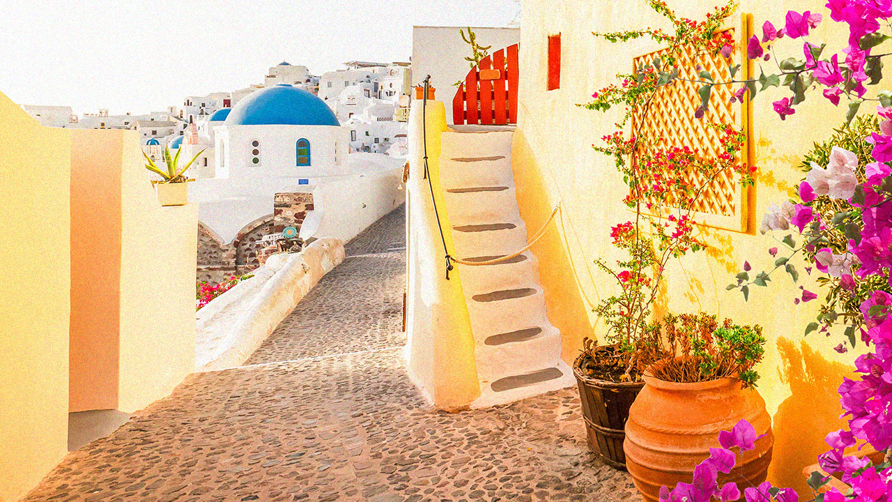 Sunlit alley in Santorini with a blue-domed building, white stairs, and vibrant pink flowers against yellow walls. Cobblestone path.