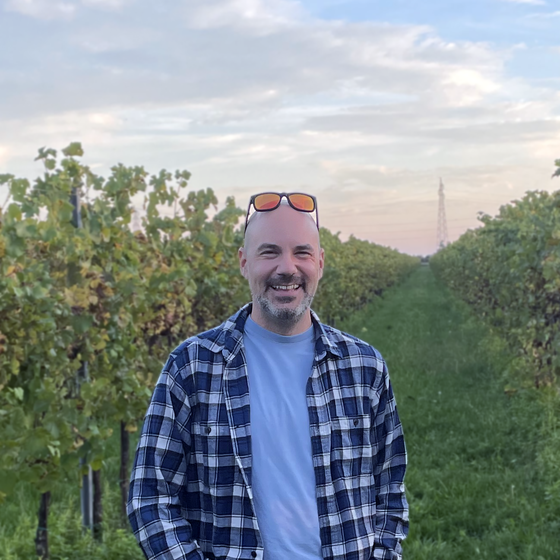 A man in a plaid shirt smiles while standing in a vineyard, with rows of grapevines and a distant power line tower under a partly cloudy sky.