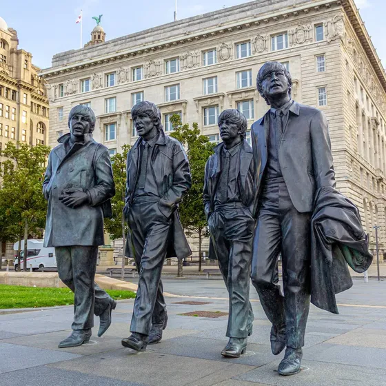 Four bronze statues of men walking, located in a plaza with historic buildings and trees in the background.