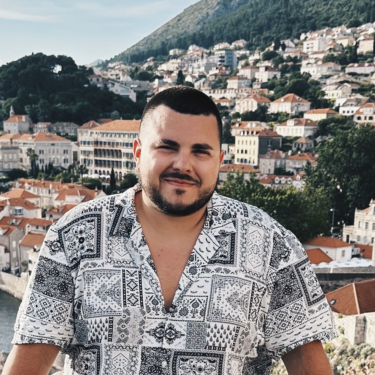 A man in a patterned shirt smiling for the camera with red-roofed buildings on a hill in the background