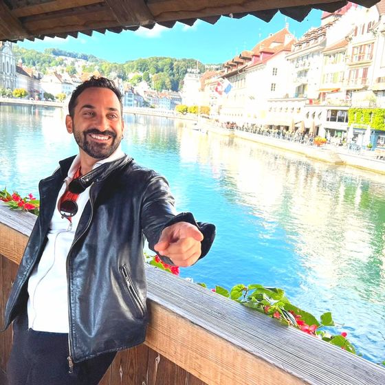 Man in a black leather jacket smiling and pointing while leaning on a railing, with a scenic riverside town and colorful houses in the background.
