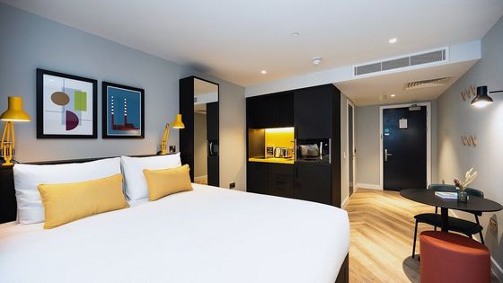 Modern hotel room with a large bed, yellow pillows, wall art, a kitchenette, round table, and a door. Natural wood flooring and warm lighting.