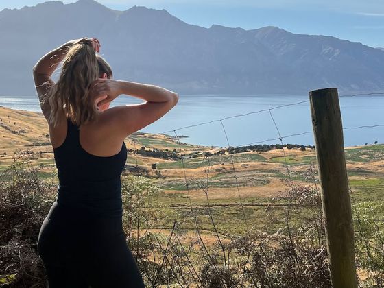 Woman in athletic wear stands by a fence, overlooking a scenic landscape with hills, a lake, and mountains in the distance.