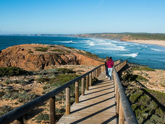 A couple walks down a wooden boardwalk toward a rugged coastline with cliffs, sandy beach, and ocean waves under a clear blue sky.