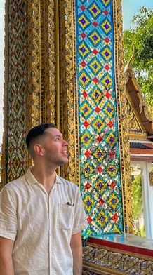 A man in a white shirt smiles while standing near colorful, ornate columns in a vibrant temple setting under a clear blue sky.