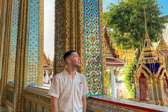 A man in a white shirt smiles while standing near colorful, ornate columns in a vibrant temple setting under a clear blue sky.