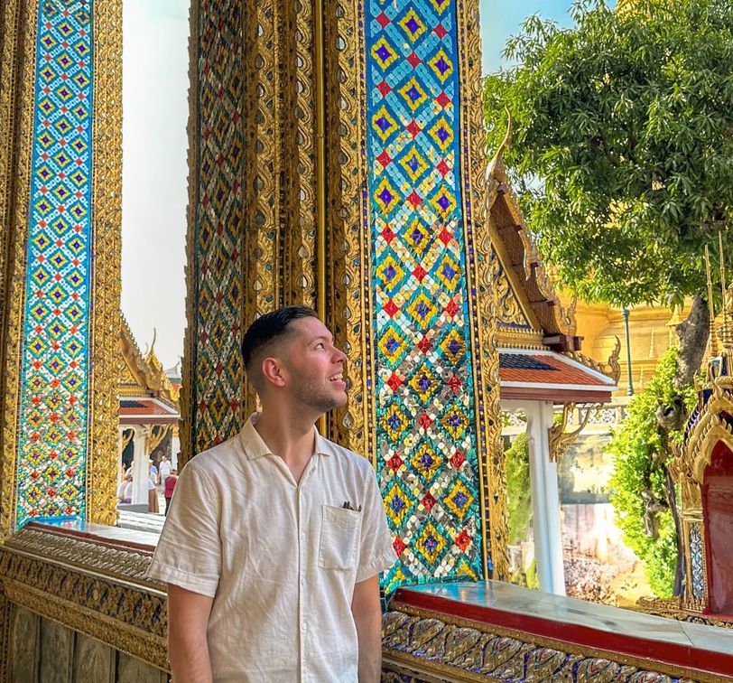 A man in a white shirt smiles while standing near colorful, ornate columns in a vibrant temple setting under a clear blue sky.