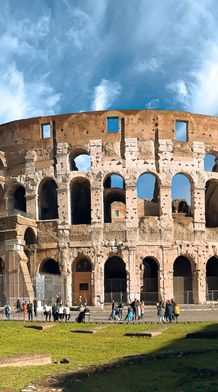 The Colosseum in Rome under a blue sky with white clouds, surrounded by tourists and green grass in the foreground.