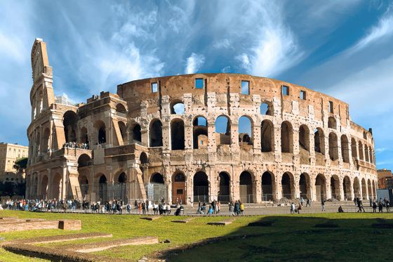 The Colosseum in Rome under a blue sky with white clouds, surrounded by tourists and green grass in the foreground.