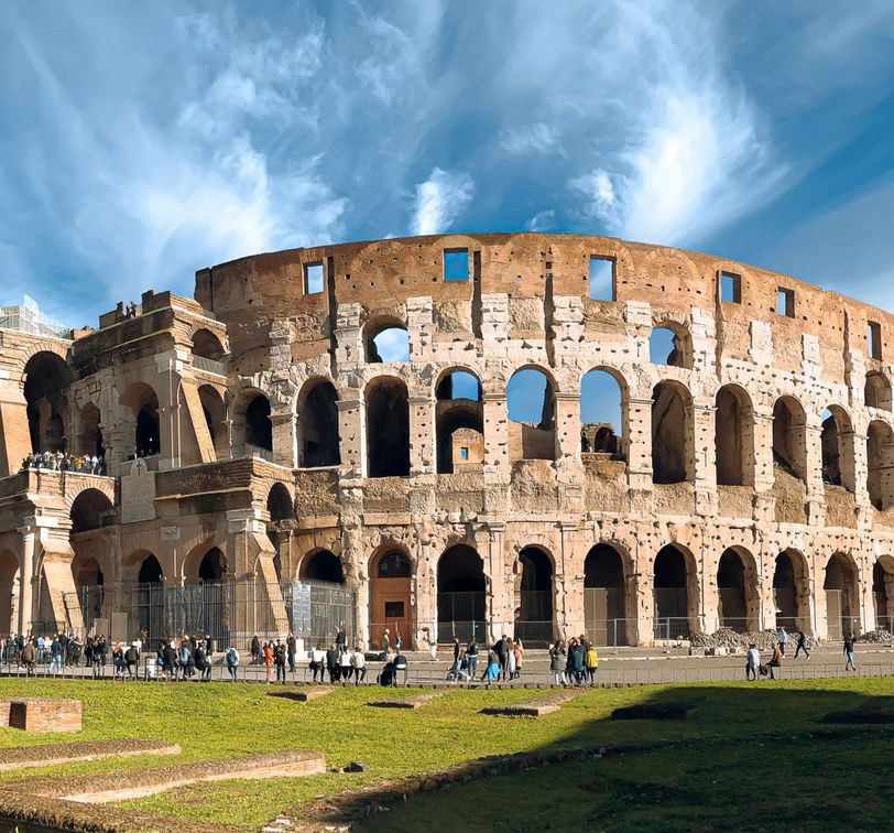 The Colosseum in Rome under a blue sky with white clouds, surrounded by tourists and green grass in the foreground.
