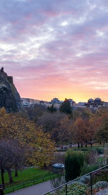 A scenic view of a castle on a hill at sunset, with a statue of a rider on horseback in the foreground, surrounded by trees and a pathway.