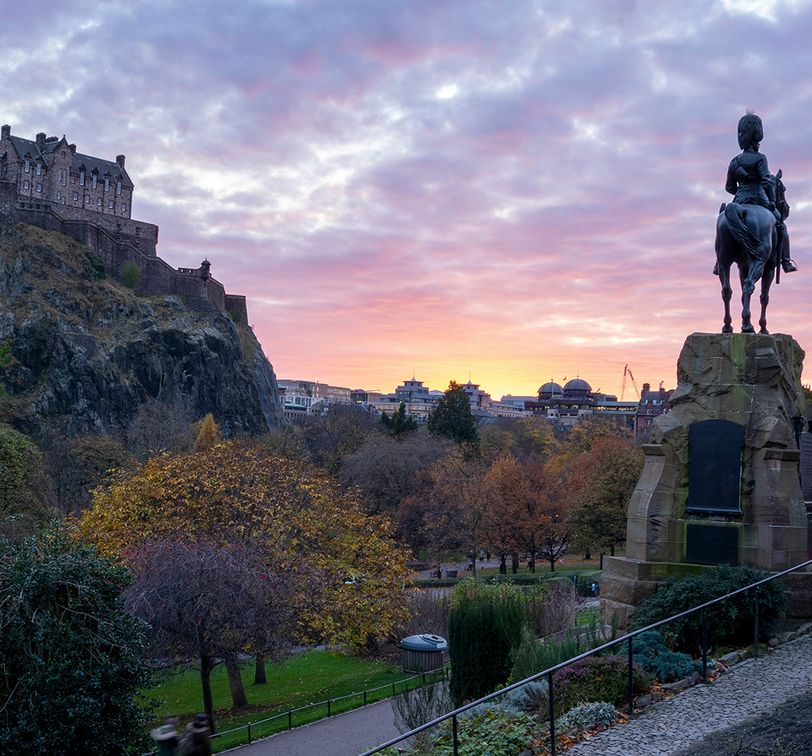 A scenic view of a castle on a hill at sunset, with a statue of a rider on horseback in the foreground, surrounded by trees and a pathway.